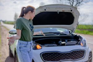 A young woman looking confused while standing next to the open hood of her car, symbolizing the common experience of hearing an unusual car noise and not knowing the cause.