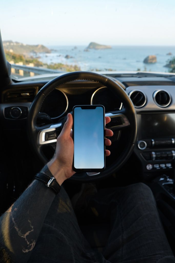 person holding cell phone behind steering wheel in car at the ocean