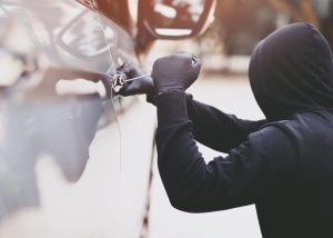 A thief uses a tool to break into a car, symbolizing the risk of vehicle theft.