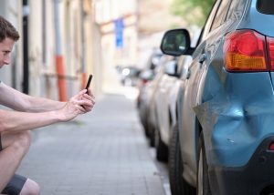 A man using his phone to take a picture of damage on his car's rear bumper, symbolizing the necessary first step of documenting damage after a hit-and-run accident.