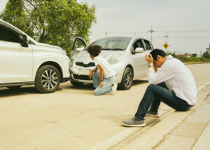 Man sitting dejected next to his severely damaged vehicle after a traffic accident, illustrating the financial and emotional fallout for uninsured drivers.